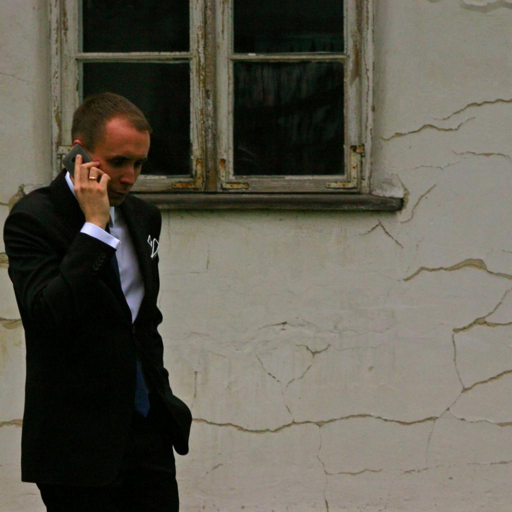 Mężczyzna w garniturze rozmawia przez telefon na tle ściany z oknem i spękaną farbą.
