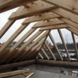 Konstrukcja więźby dachowej z desek obrzynanych, widok od wewnątrz, zarys zabudowy w tle.