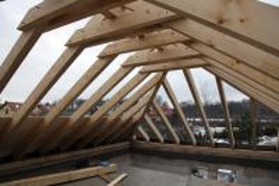 Konstrukcja więźby dachowej z desek obrzynanych, widok od wewnątrz, zarys zabudowy w tle.