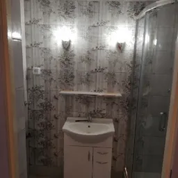 Ukończona łazienka z kabiną prysznicową, umywalką z szafką i płytkami ściennymi w kwiatowy wzór, oświetlona dwoma kinkietami.