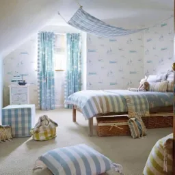 Pokój dziecięcy w stylu marynistycznym: białe ściany z tapetą w żaglówki, niebiesko-białe zasłony i baldachim nad łóżkiem, drewniane drzwi z mosiężną klamką w prawym rogu.