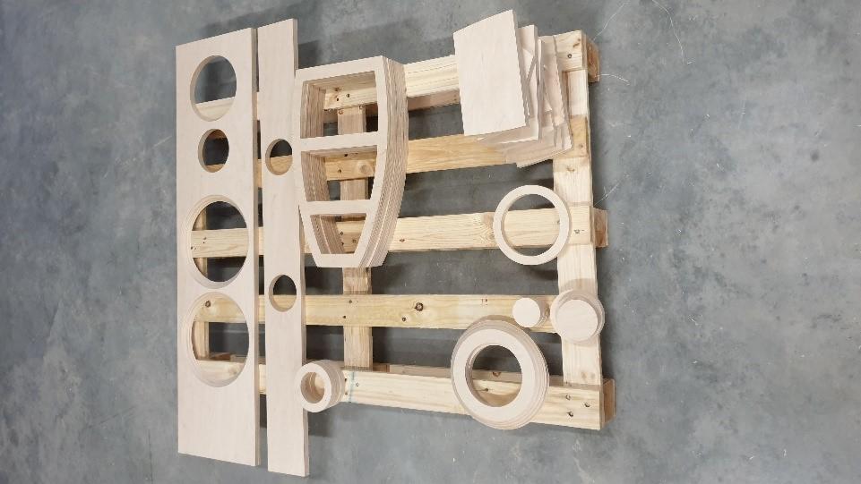 Elementy z jasnego drewna, wycięte i ułożone na drewnianej palecie, niektóre z okrągłymi otworami, prawdopodobnie przygotowane do montażu konstrukcji.