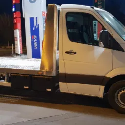 Biała laweta z płaską platformą stoi na parkingu stacji benzynowej Verva, widoczny bok pojazdu i fragment platformy załadunkowej.