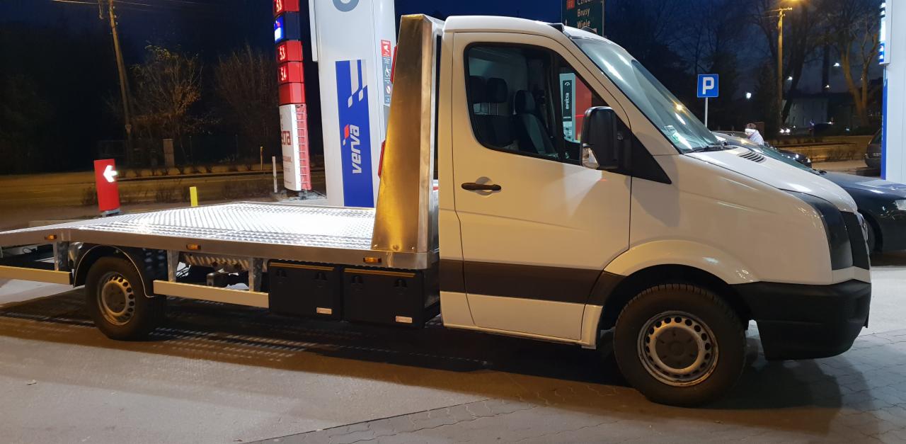 Biała laweta z płaską platformą stoi na parkingu stacji benzynowej Verva, widoczny bok pojazdu i fragment platformy załadunkowej.