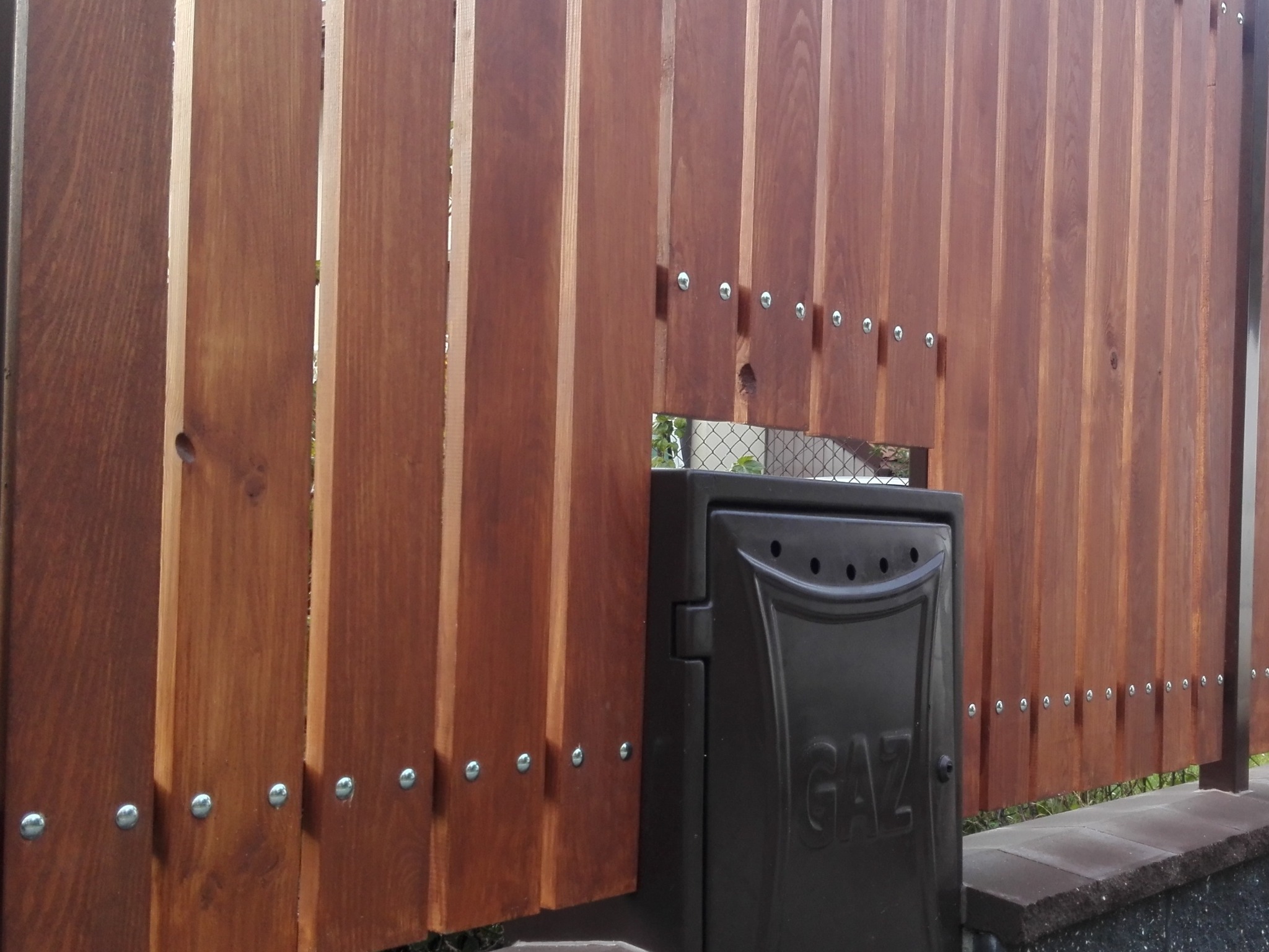 Nowoczesne ogrodzenie z drewnianych desek w odcieniach brązu, przymocowanych za pomocą srebrnych śrub, z czarną skrzynką gazową z napisem 'GAZ' w dolnym rogu.