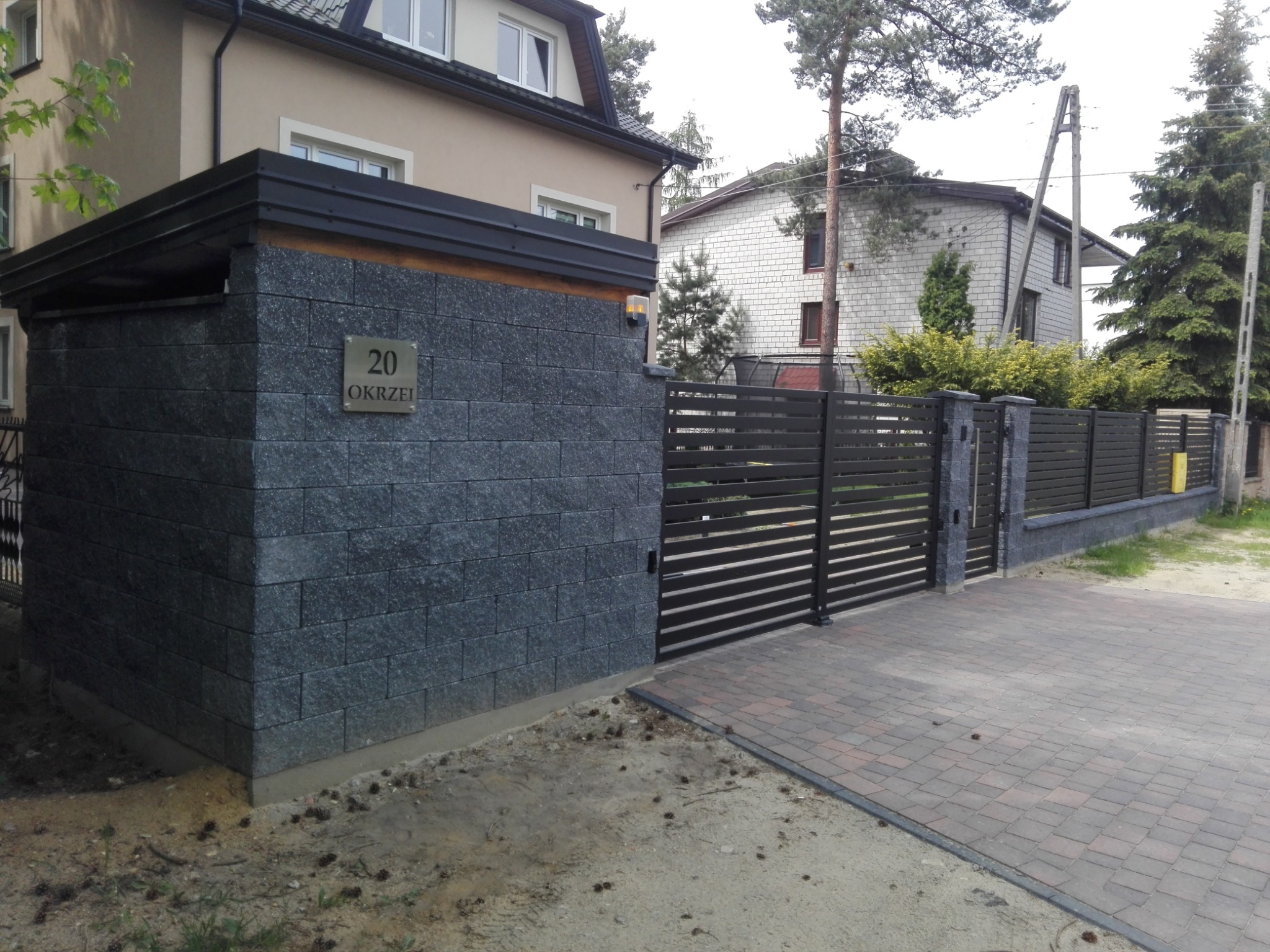 Nowoczesne ogrodzenie posesji z czarną, poziomą bramą i szarym, kamiennym słupkiem z numerem domu '20 Okrzei' w słoneczny dzień.