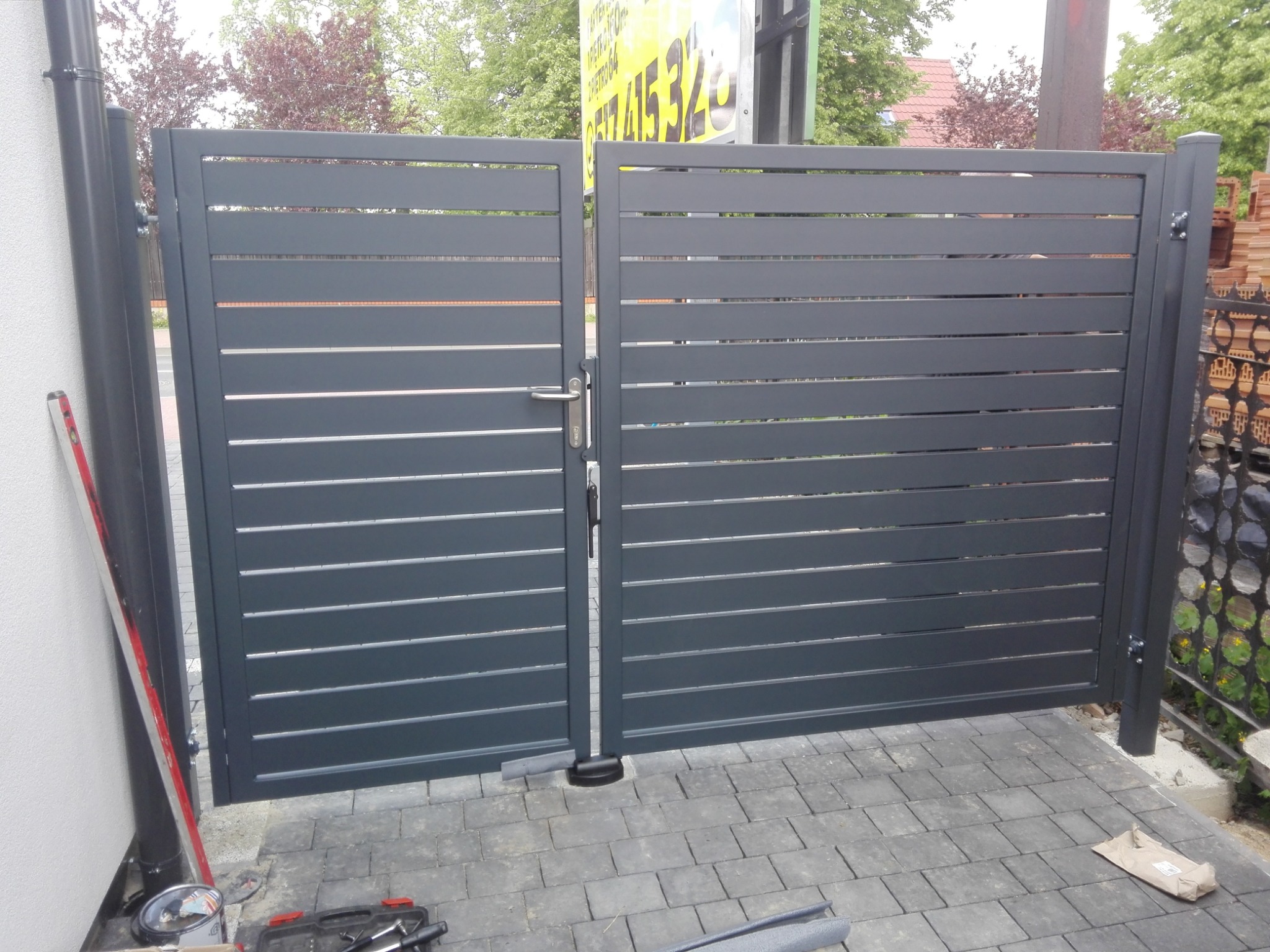 Nowoczesna, grafitowa brama posesyjna z poziomymi panelami, częściowo otwarta, widoczna skrzynka z narzędziami i poziomica przy słupku, kostka brukowa na podjeździe.