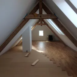 Montaż drewnianej podłogi na poddaszu ze skośnymi ścianami i widoczną konstrukcją więźby dachowej; częściowo ułożone panele na brązowej płycie podkładowej, w tle okno i wyposażenie.