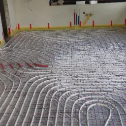 Rozłożona instalacja ogrzewania podłogowego na folii izolacyjnej z siatką, rury zabezpieczone taśmą przy ścianach, widoczne fragmenty instalacji wodnej w ścianie.