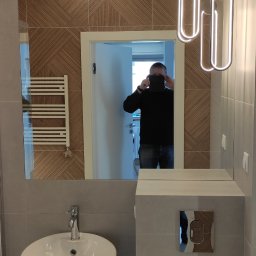 OPALIŃSKI Nowoczesne Wykończenia - Nowoczesna łazienka z kabiną prysznicową, umywalką nablatową i grzejnikiem drabinkowym, wykończona płytkami w odcieniach szarości i drewna, z odbiciem osoby robiącej zdjęcie w lustrze.