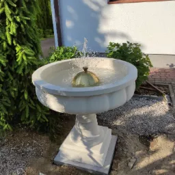 Biała, betonowa fontanna ogrodowa z złotą kulą w środku, tryskająca wodą, otoczona zielenią, na tle białego domu z brązowymi ramami okiennymi.