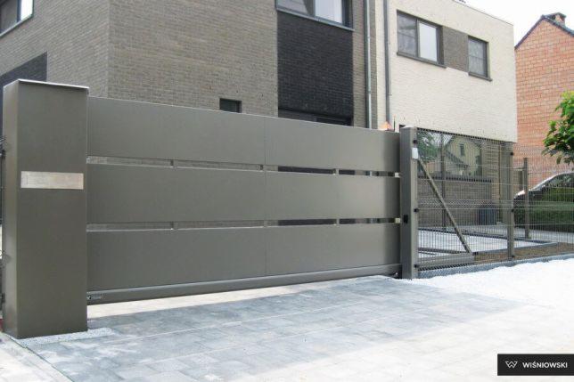 Szara, przesuwna brama wjazdowa z poziomymi panelami, zamontowana przed nowoczesnym domem z cegły, widoczna kostka brukowa na podjeździe.