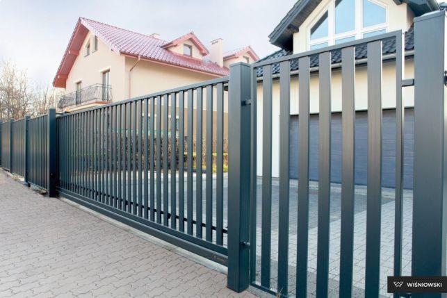 Nowoczesna, przesuwna, grafitowa brama wjazdowa z pionowymi szczeblami, z widocznym fragmentem domu z garażem w tle.