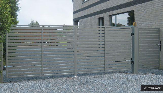 Nowoczesna, pozioma brama wjazdowa z furtką, szara, z widocznym logo producenta Wiśniowski w prawym dolnym rogu, na tle domu z cegły.