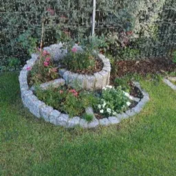 Spiralna rabata kwiatowa z kostki granitowej, obsadzona różami i innymi kwiatami, na tle trawnika i siatki ogrodzeniowej.