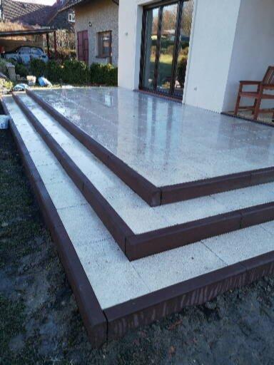 Nowoczesny taras z jasnoszarej kostki brukowej wykończony ciemnobrązową obwódką, prowadzący do przeszklonych drzwi tarasowych domu jednorodzinnego.