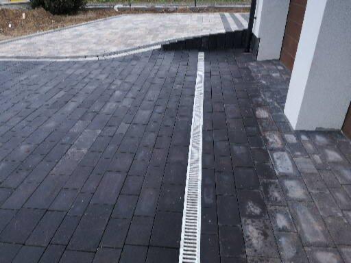 Ułożona ciemna kostka brukowa z odpływem liniowym na podjeździe, widoczne ślady po wodzie na kostce, krawężnik z jasnej kostki w tle.