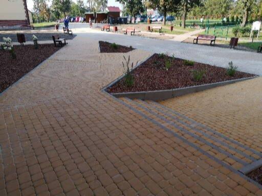 Ułożona kostka brukowa w kolorze piaskowym tworząca ścieżkę i schody, z elementami dekoracyjnymi z brązowego grysu i zieleni w kształcie trójkątów, na tle parku z ławkami.