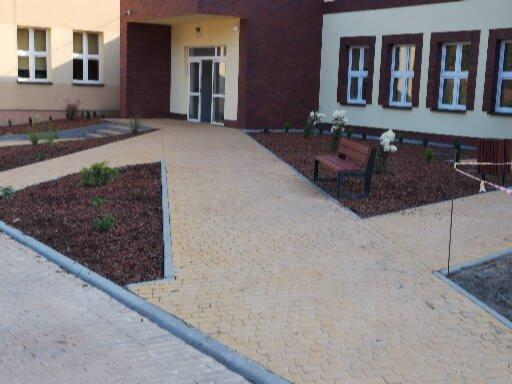 Żółta kostka brukowa ułożona w ścieżkę przed budynkiem, z rabatami wysypanymi korą i niskimi krzewami po bokach, ławka w tle.