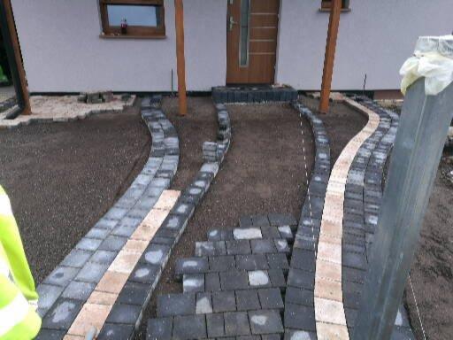 Fragment układanej ścieżki z kostki brukowej w kolorach szarości i beżu, prowadzącej do drzwi wejściowych domu, widoczne nierówności terenu i niedokończone etapy prac.