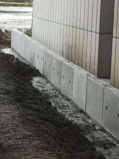 Świeżo ułożony betonowy obrzeżnik chodnikowy z otworami drenażowymi, przylegający do ściany budynku, widoczny mokry grunt po pracach.