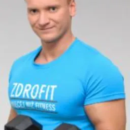 Mężczyzna w błękitnej koszulce Zdrofit, z hantlą w prawej ręce, uśmiecha się do kamery na jasnoszarym tle. Widoczny biceps i krótko ostrzyżone włosy.