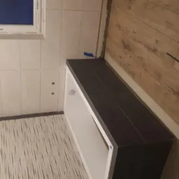 Wnętrze łazienki w trakcie remontu: białe płytki ścienne, podłoga z wzorzystych płytek, zabudowa pod umywalkę z ciemnymi płytkami oraz fragment podłogi z paneli drewnopodobnych, widoczne narzędzia...