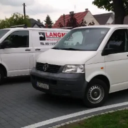 Dwa białe samochody dostawcze firmy instalacyjnej Langner z widocznym logo i numerami telefon&oacute;w, zaparkowane na brukowanym podjeździe przed budynkiem mieszkalnym.
