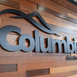 Two Squares Project - Metalowe logo 'Columbia distributing' na drewnianej ścianie z poziomych paneli w różnych odcieniach brązu.