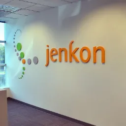 Pomarańczowe, przestrzenne litery z nazwą firmy 'jenkon' i graficznym elementem z kolorowych kropek na białej ścianie w biurze z widokiem na zewnątrz.