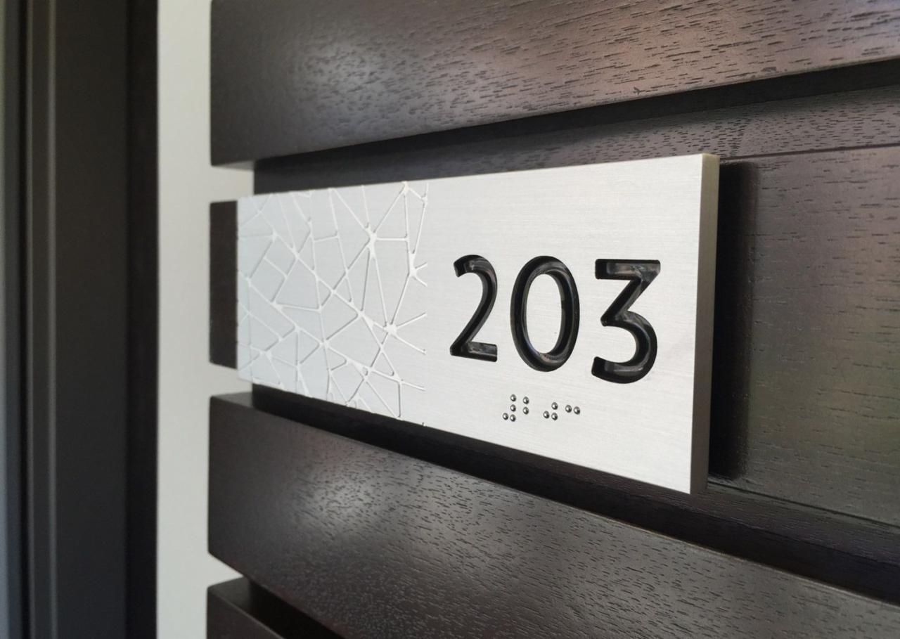 Metalowa tabliczka z numerem 203 i wypukłym wzorem geometrycznym, przymocowana do ciemnego, drewnianego panelu drzwiowego, zawiera oznaczenia w alfabecie Braille'a.