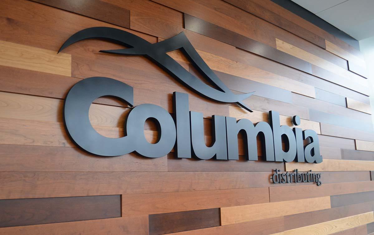Metalowe logo 'Columbia distributing' na drewnianej ścianie z poziomych paneli w różnych odcieniach brązu.