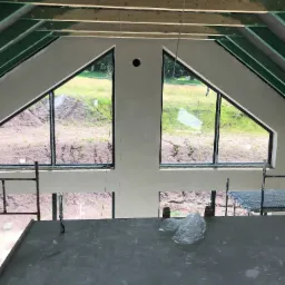 Wnętrze w trakcie prac wykończeniowych: surowa posadzka, świeżo otynkowana ściana z dwoma trójkątnymi oknami z widokiem na zewnątrz, widoczne elementy konstrukcji drewnianej dachu.