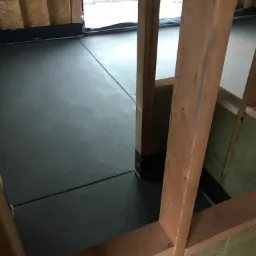 Szara, gładka wylewka podłogowa w surowym wnętrzu, widoczna przez drewnianą konstrukcję szkieletową, z izolacją z wełny mineralnej przy ścianach.