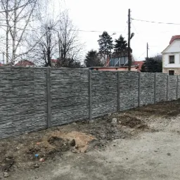 Nowo postawiony, szary, betonowy płot imitujący kamień, z widocznymi słupkami, na tle ziemi i budynków mieszkalnych w pochmurny dzień.