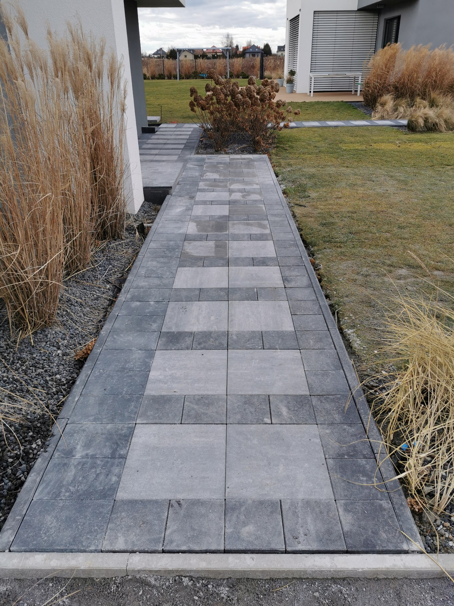 Ułożona ścieżka z szarej kostki brukowej prowadząca przez ogród z ozdobnymi trawami i krzewami, widok z perspektywy pieszego.
