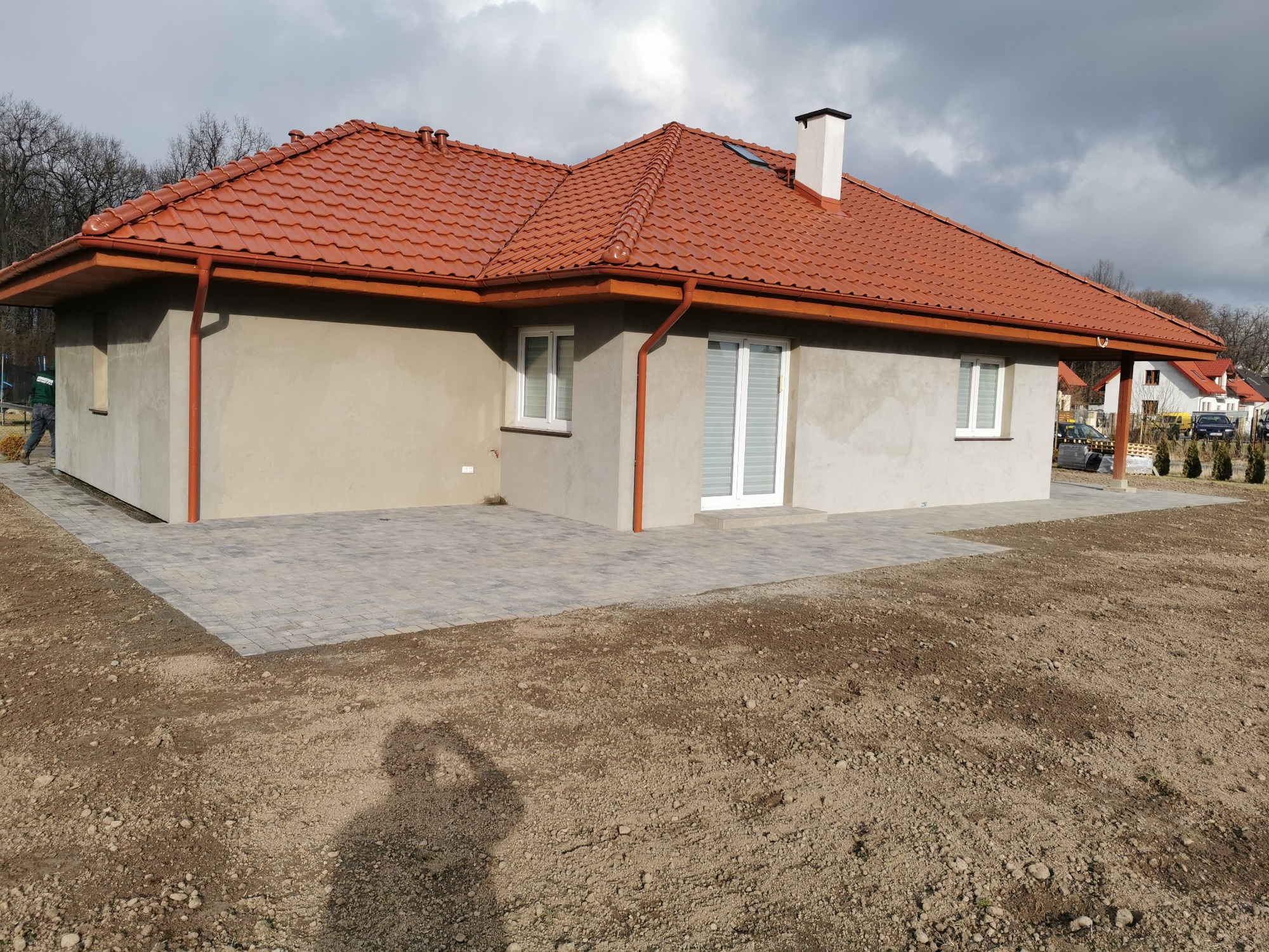 Nowo wybudowany dom jednorodzinny z czerwoną dachówką i szarą elewacją, otoczony świeżo ułożoną kostką brukową i przygotowanym terenem pod trawnik, widoczne okna z białymi ramami i biały komin.