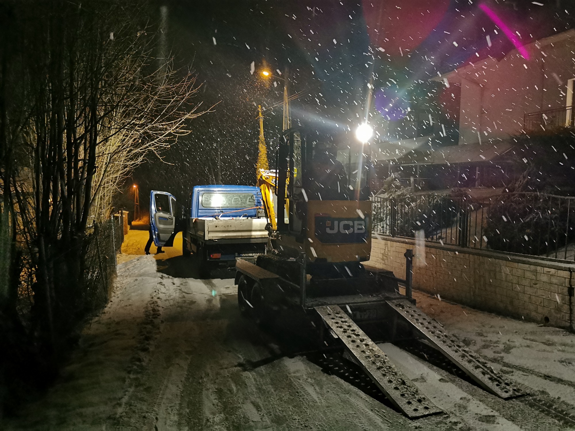 Nocne zdjęcie zaśnieżonej ulicy z widocznym samochodem dostawczym z otwartymi drzwiami i przyczepą z koparką JCB, oświetlone latarnią i światłami pojazdów, w tle budynek.