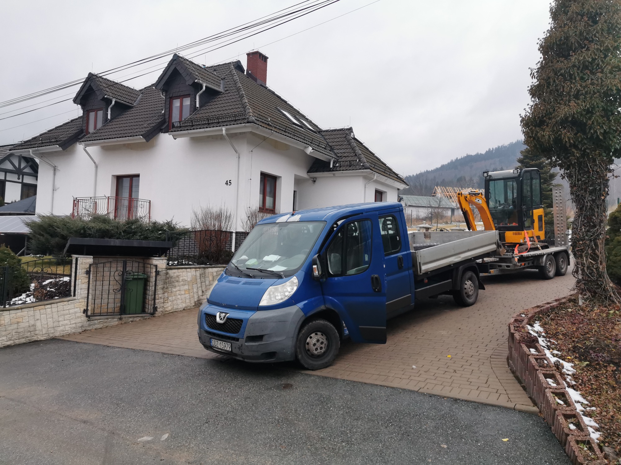 Niebieski samochód dostawczy z otwartymi drzwiami, przewożący małą koparkę na przyczepie, zaparkowany na kostce brukowej przed białym domem z ciemnym dachem, w tle zalesione wzgórze.