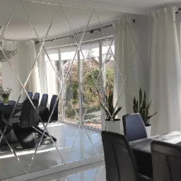 Salon z lustrzaną ścianą w romby, odbijającą stół z czarnymi krzesłami i widok na ogród za oknem.