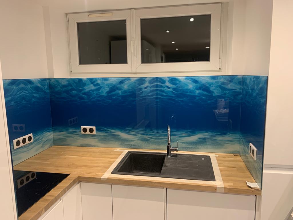 Szklana ściana nad blatem kuchennym z motywem podwodnego krajobrazu, widoczne gniazdka elektryczne i okno w tle.