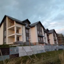 Nowe, dwukondygnacyjne budynki mieszkalne z balkonami, pomalowane na biało i szaro, widoczne zza metalowego ogrodzenia i wysokiej trawy, z budynkiem w trakcie budowy w tle.