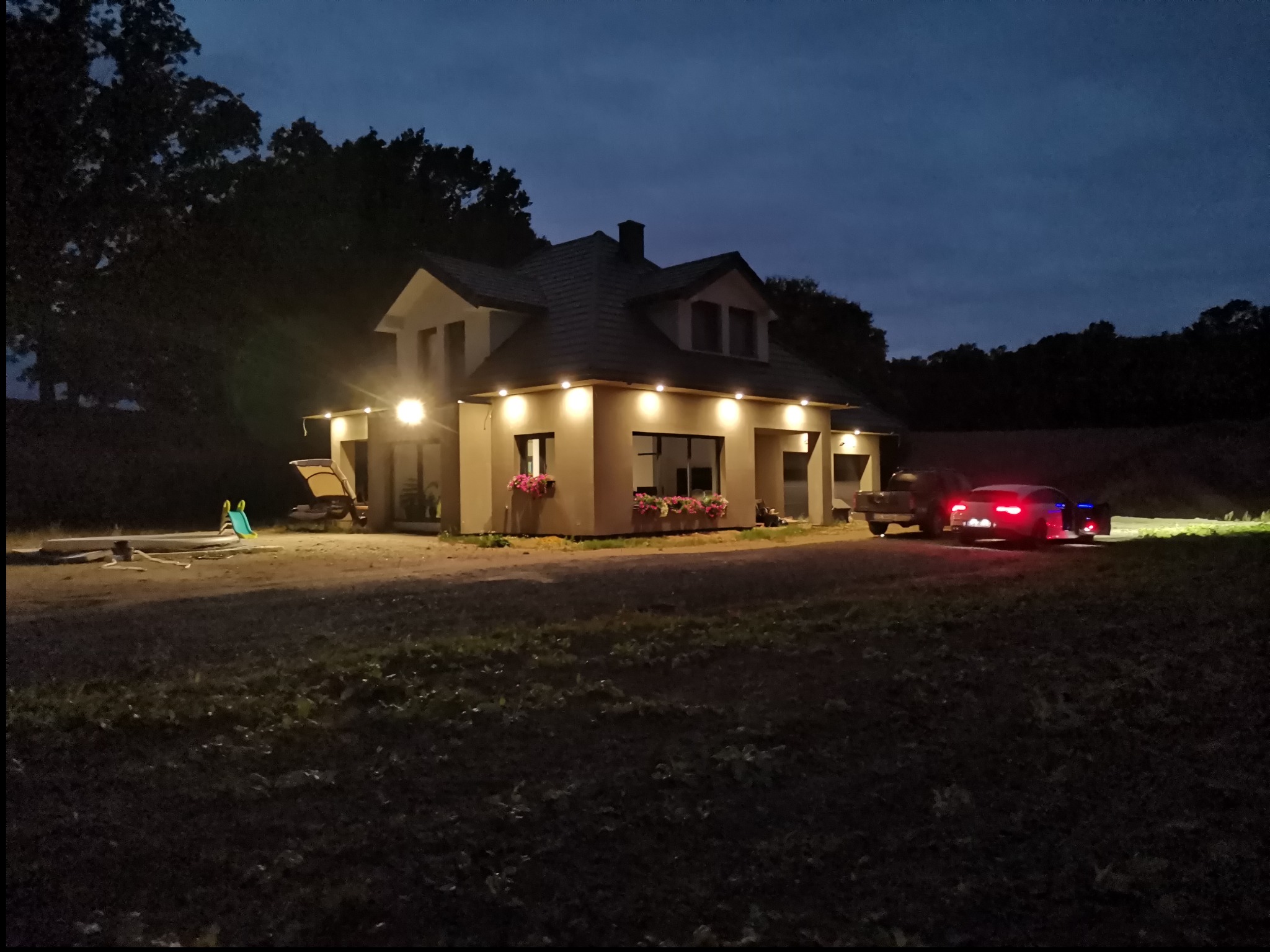 Wieczorny widok na dom jednorodzinny z oświetleniem elewacyjnym LED, widoczne światła samochodu w tle, trawnik i drzewa.