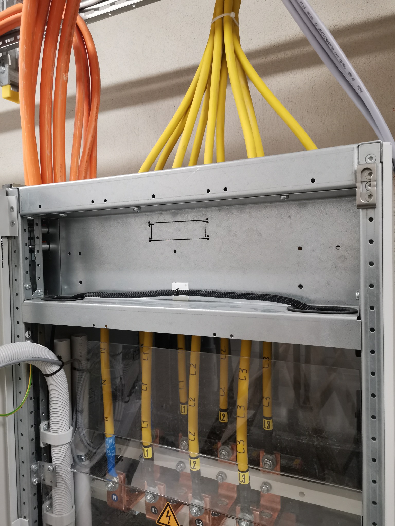 Szczegółowe ujęcie rozdzielnicy elektrycznej z widocznymi kablami oznaczonymi L1, L2, L3 oraz N, zabezpieczonymi przezroczystą osłoną i z symbolem ostrzegawczym.