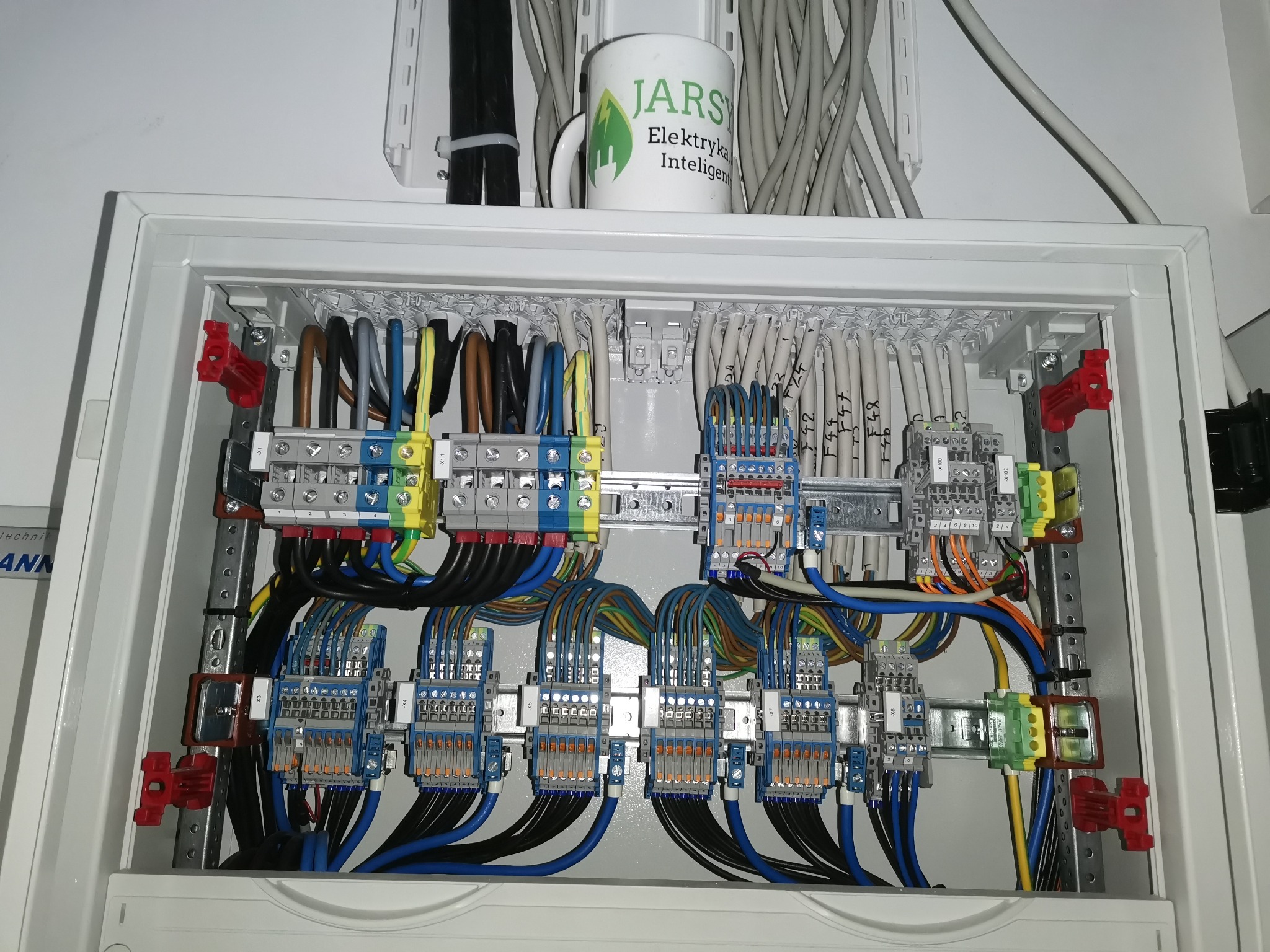 Otwarta skrzynka rozdzielcza z licznymi przewodami, listwami zaciskowymi i bezpiecznikami; na górze kubek z logo firmy elektrycznej.