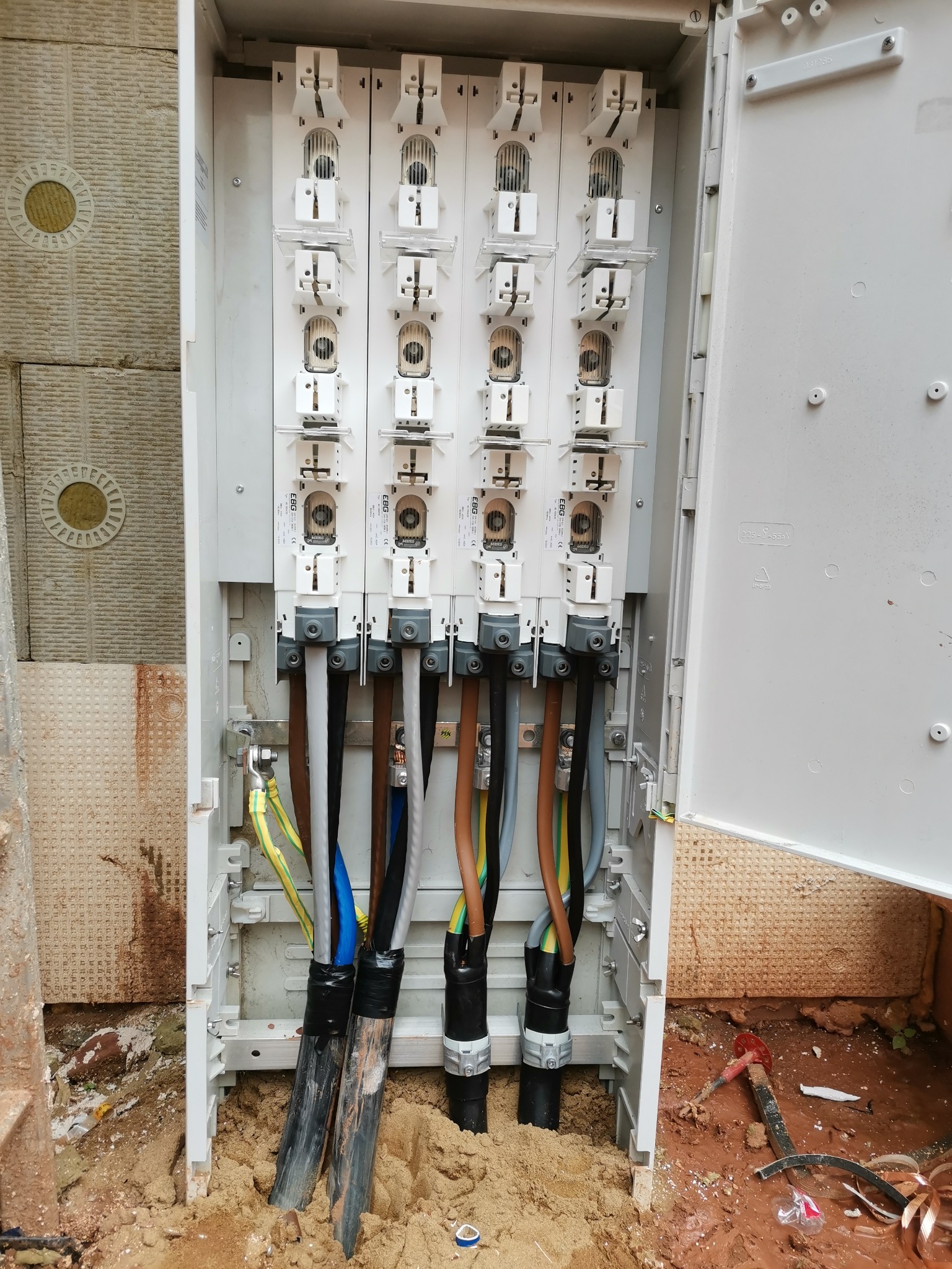 Otwarta skrzynka rozdzielcza z podłączonymi grubymi kablami elektrycznymi o różnych kolorach, zamocowanymi w białych uchwytach, widoczna ściana budynku z ociepleniem.