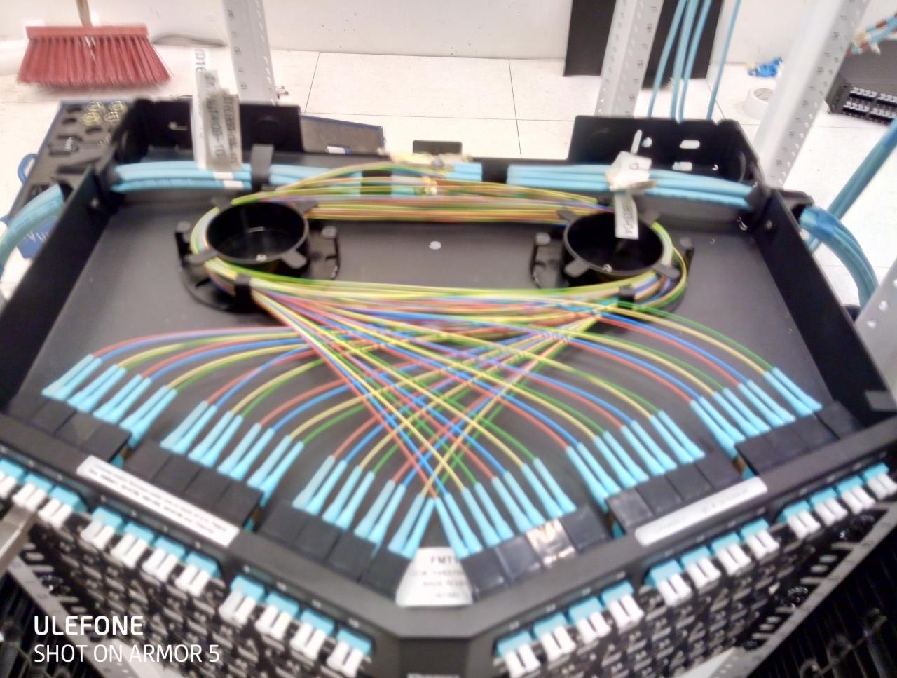 Rozdzielnica światłowodowa z wielokolorowymi kablami i złączami, widok z góry, z elementami infrastruktury telekomunikacyjnej w tle.