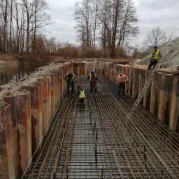 Budowa fundamentów: Widok z góry na przygotowany szalunek z metalowych ścianek i zbrojenie z siatki stalowej, kilku robotników w kaskach i kamizelkach odblaskowych na placu budowy, drabina oparta...