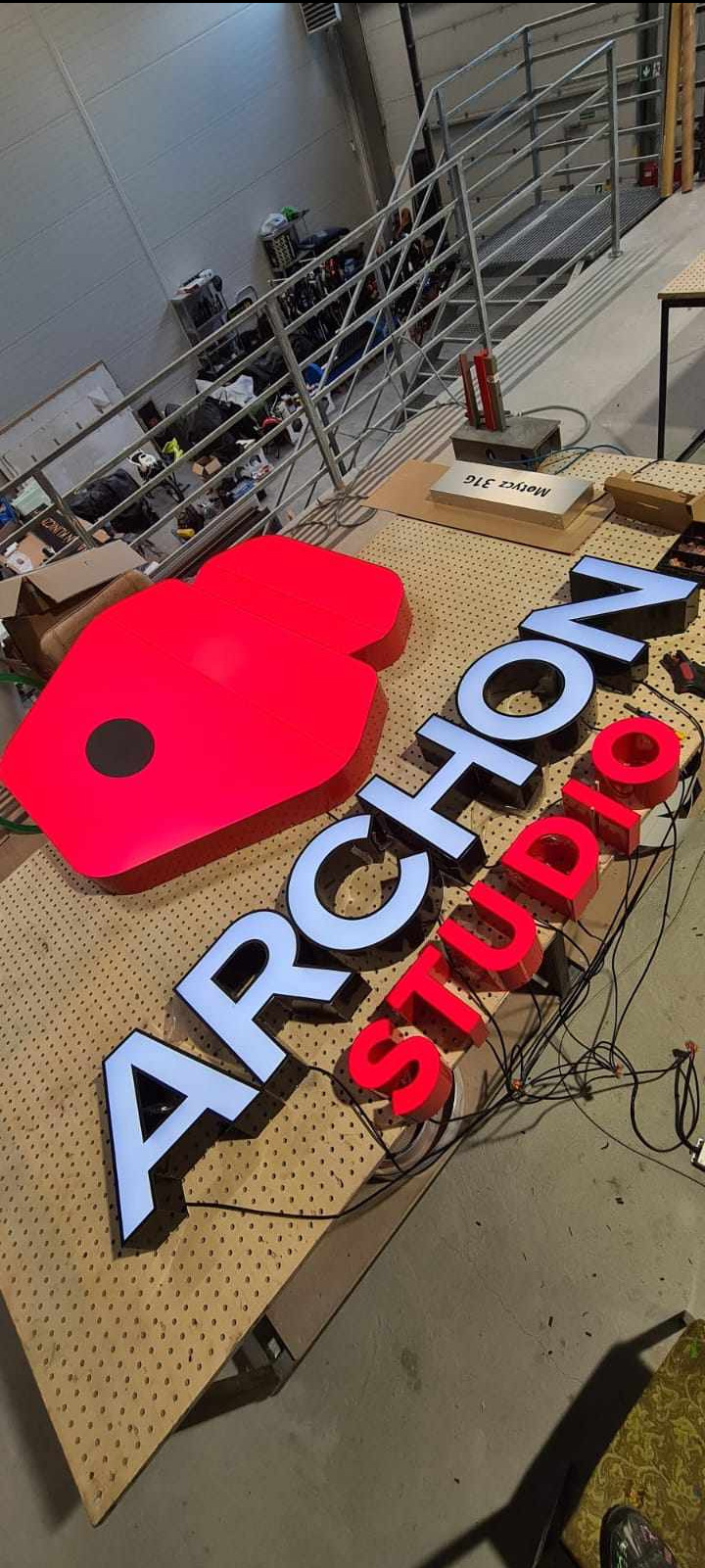 Niedokończona instalacja podświetlanego logo 3D 'Archon Studio' z czerwonymi i białymi elementami na perforowanym stole warsztatowym, widoczne kable i zasilacze.