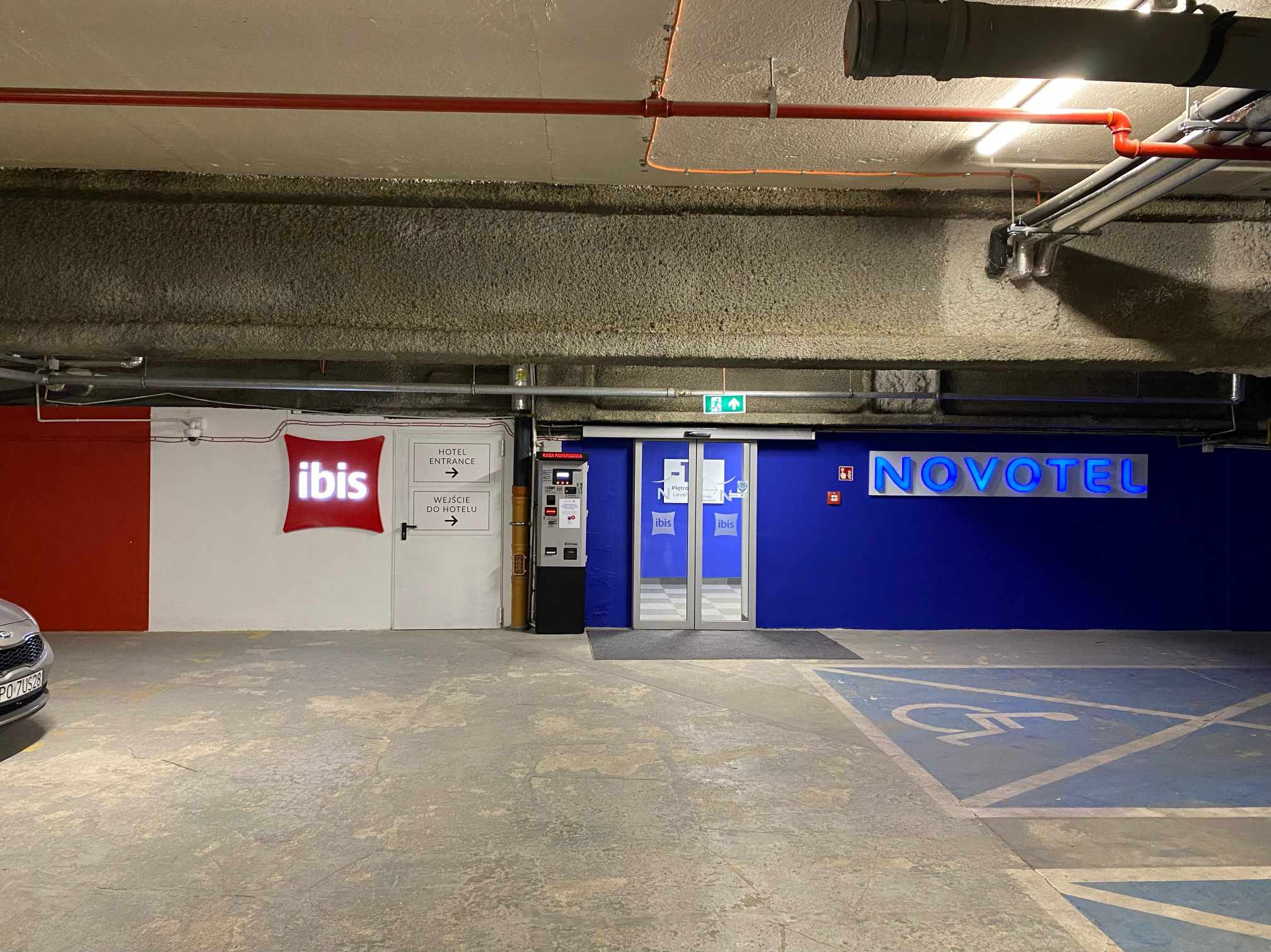Wnętrze parkingu podziemnego z widocznymi oznaczeniami hoteli Ibis i Novotel, automat parkingowy i strzałki wskazujące kierunek wejścia do hotelu.